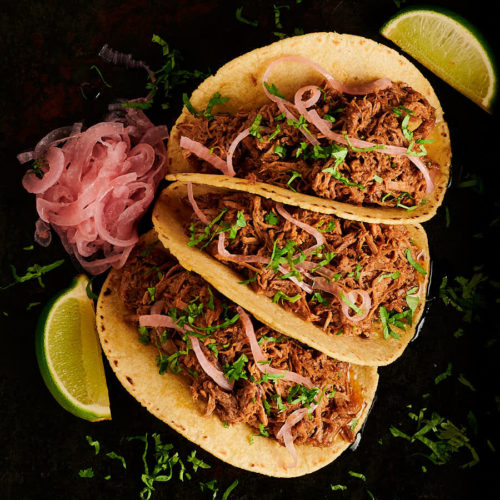 Beef barbacoa tacos with pickled onions and lime wedges from above.