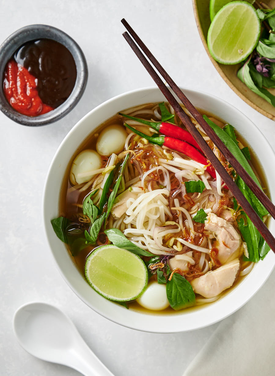 Bowl of pho ga with chopsticks with hoisin and sriracha side dishes.