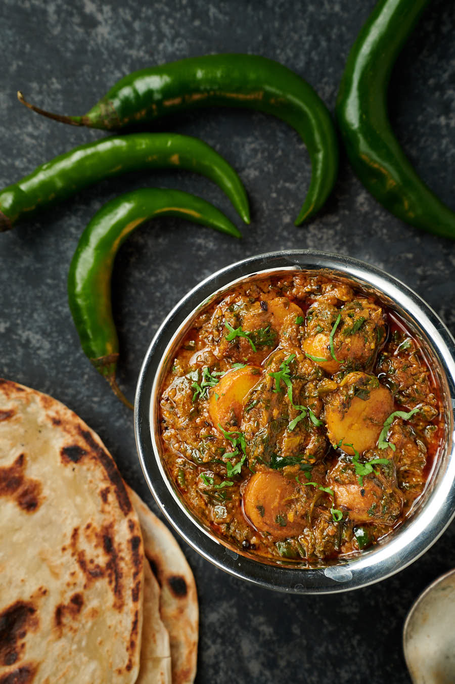 Saag aloo in a bowl surrounded by green chilies and parathas.