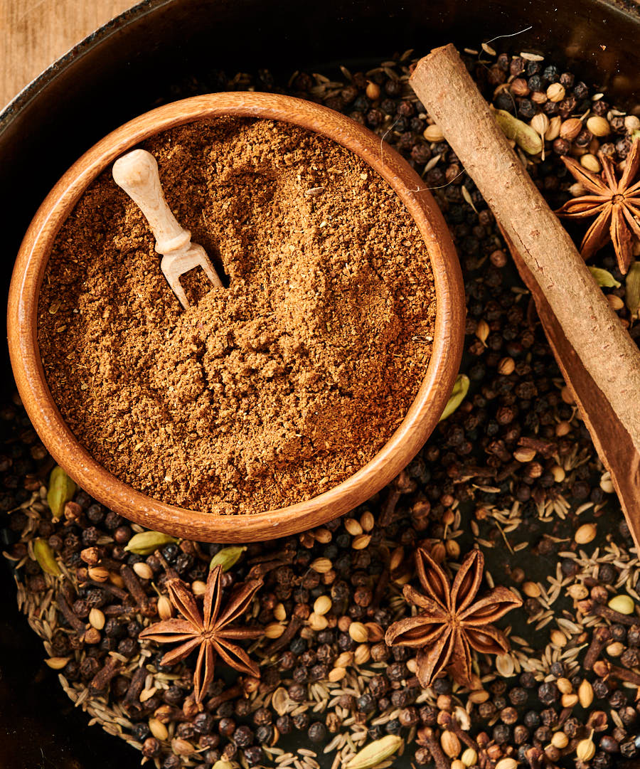 Bowl of garam masala surrounded by peppercorns, cumin seed, cinnamon and star anise from above.