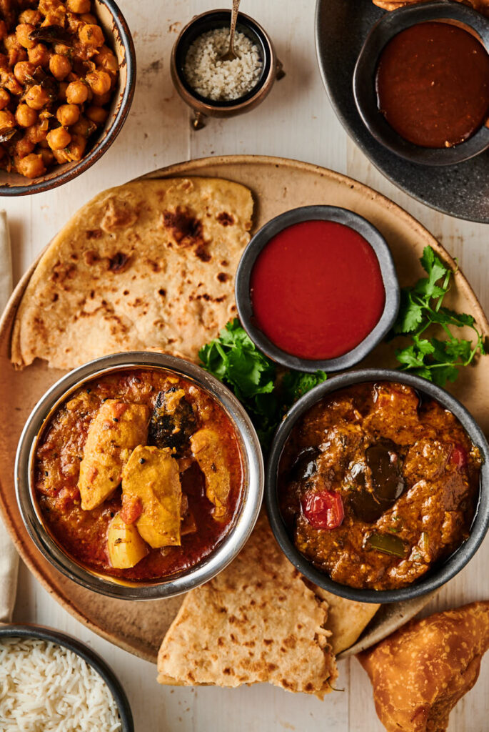 Table scene from above. Murgir jhol, eggplant curry, chili sauce, parathas, chana masala and samosas. From above.