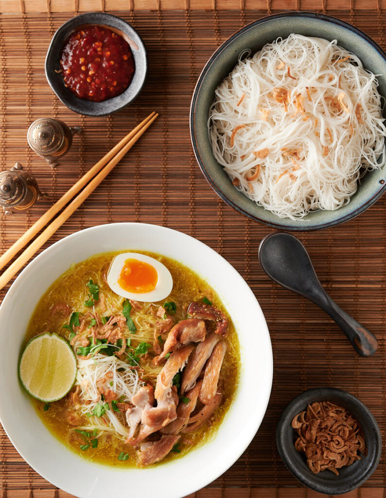 Table scene with soto ayam, rice noodles, dried shallots and sambal oelek.