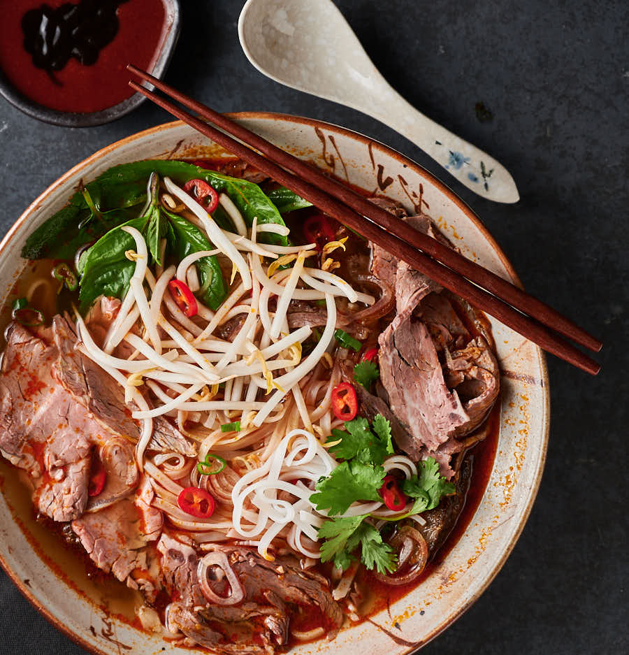 Table scene of spicy beef noodle soup, spoon and hoisin sriracha dipping bowl.