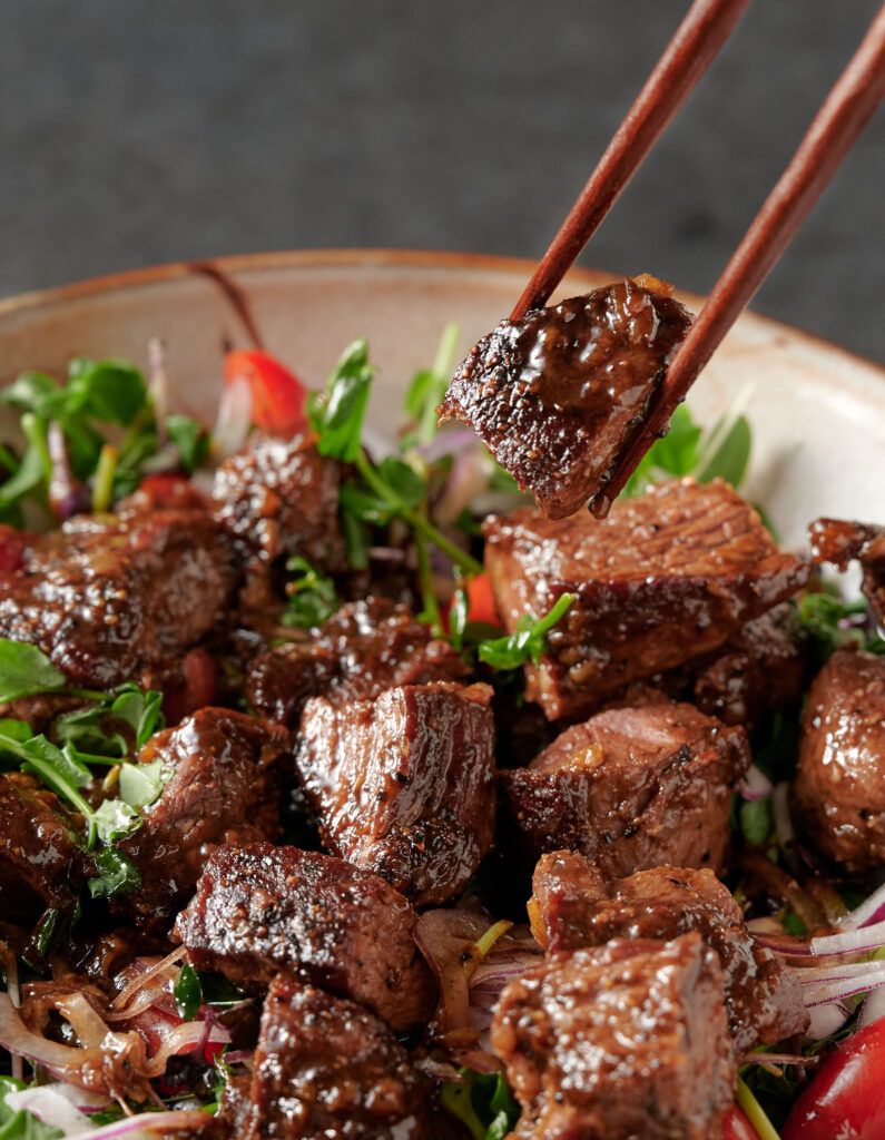 Piece of shaking beef held in chopsticks above a closeup of bo luc lac.