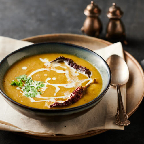 Bowl of mulligatawny soup garnished with drizzled cream, cilatntro and whole kashmiri chilies from the front.