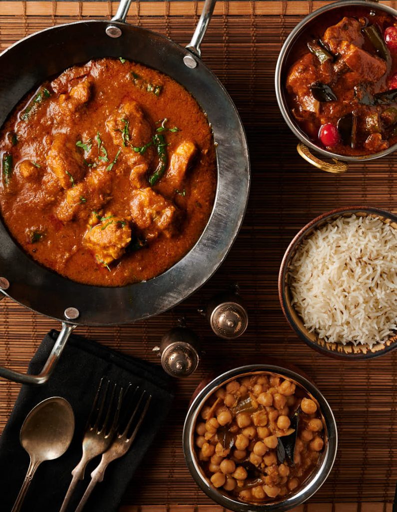 Table scene featuring achari chicken in a kadai surrounded by smaller side dishes.