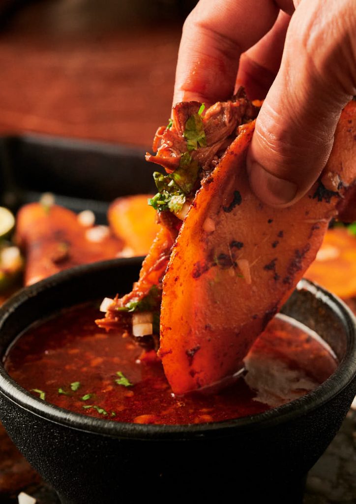 Hand holding birria taco dipped into consomé from the front.