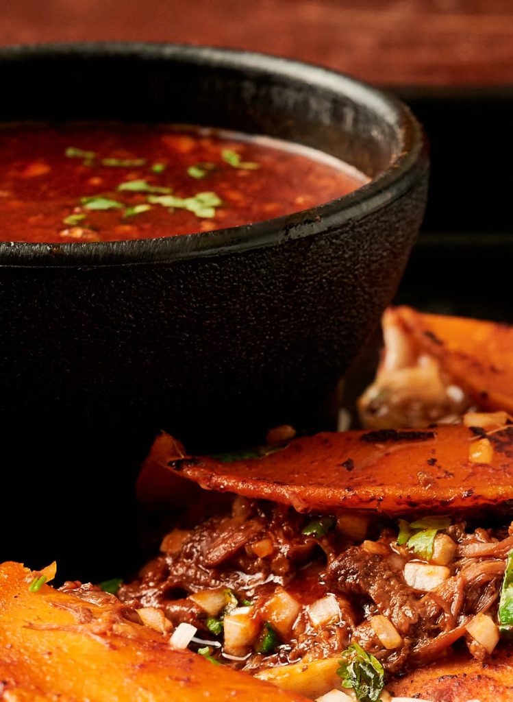 Bowl of birria de res consomé surrounded by birria tacos from the front.