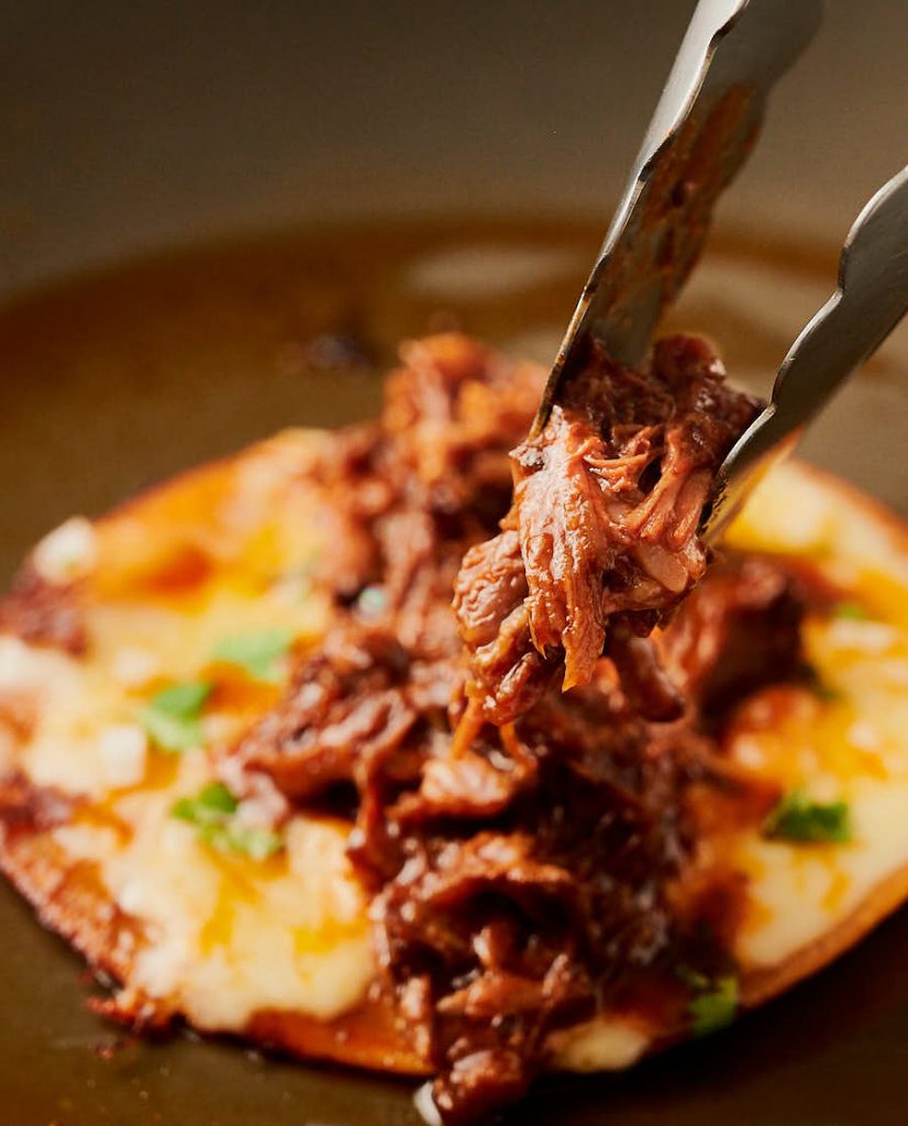 Birria being spread out on a cheesy fried tortilla base.