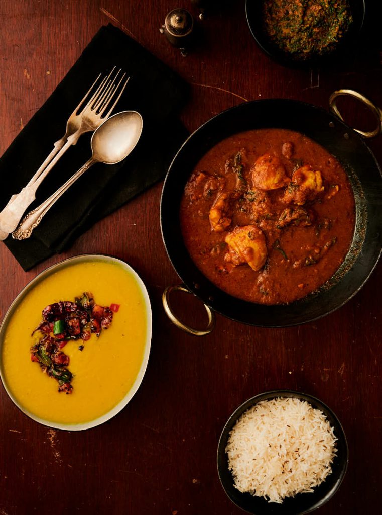 Table scene with hotel style methi chicken in a kadai with a dal tarka, rice and spinach curry.