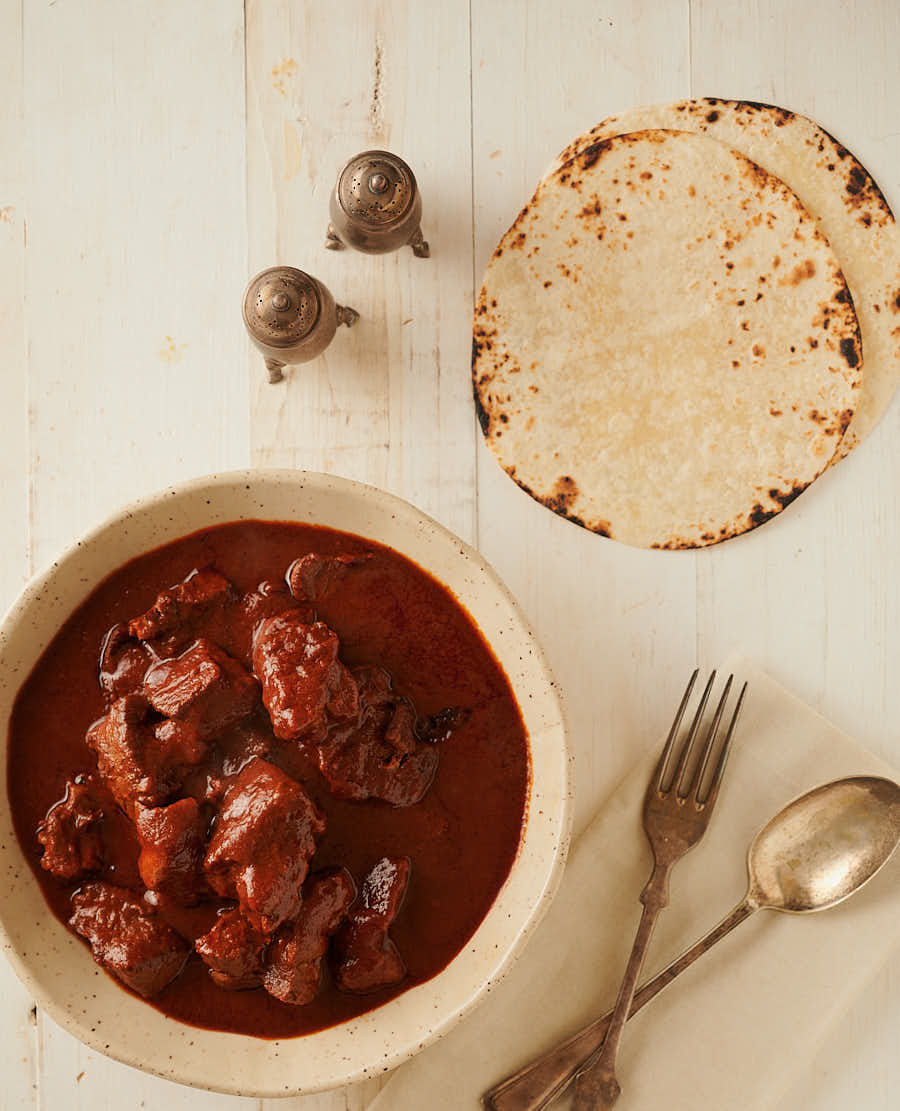 Table scene with bowl of carne adovada, tortillas and cutlery from above.